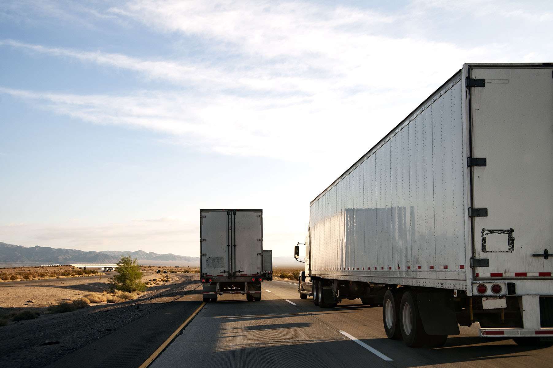 Multiple white semi-trucks drive up a road in the desert.
