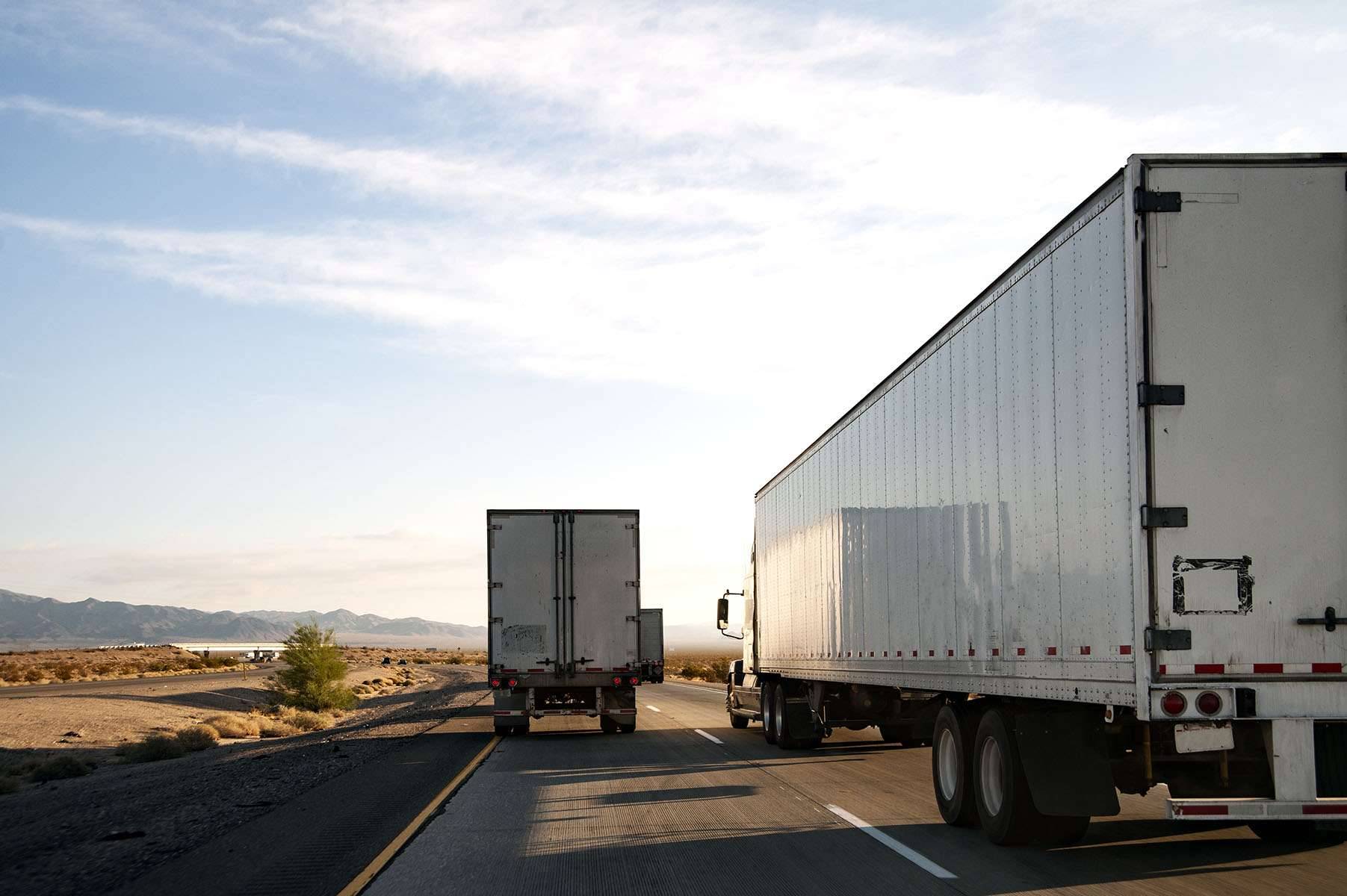 Multiple white semi-trucks drive up a road in the desert.