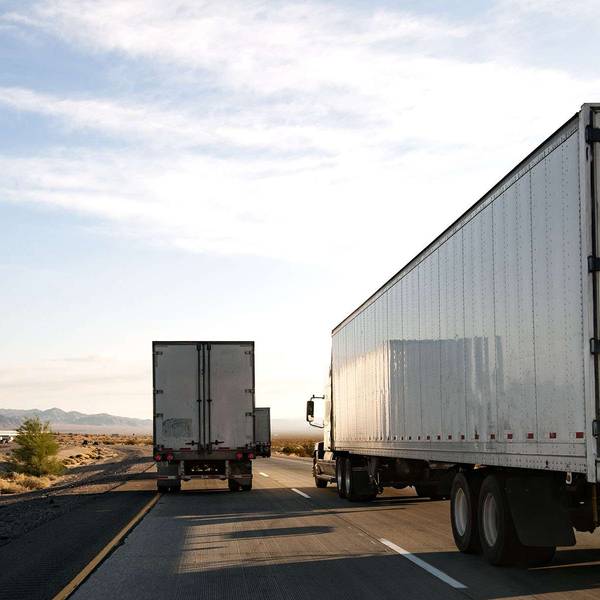 Multiple white semi-trucks drive up a road in the desert.