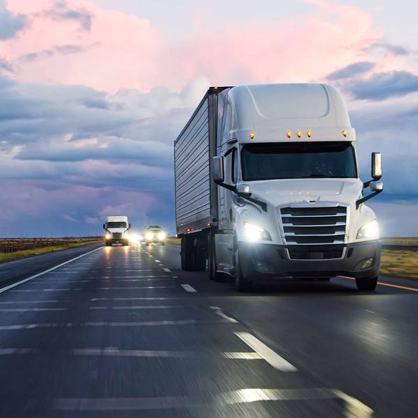 Several white semi-trucks drive at sunset on a highway.