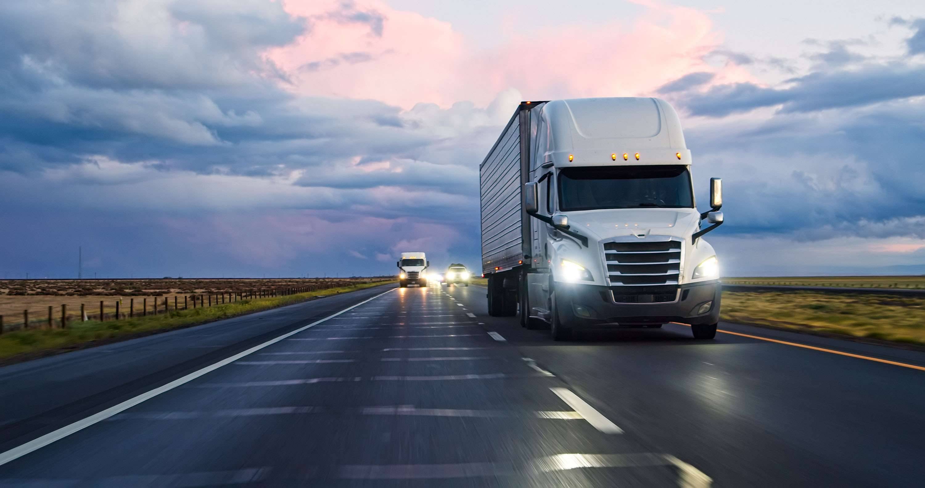 Several white semi-trucks drive at sunset on a highway.