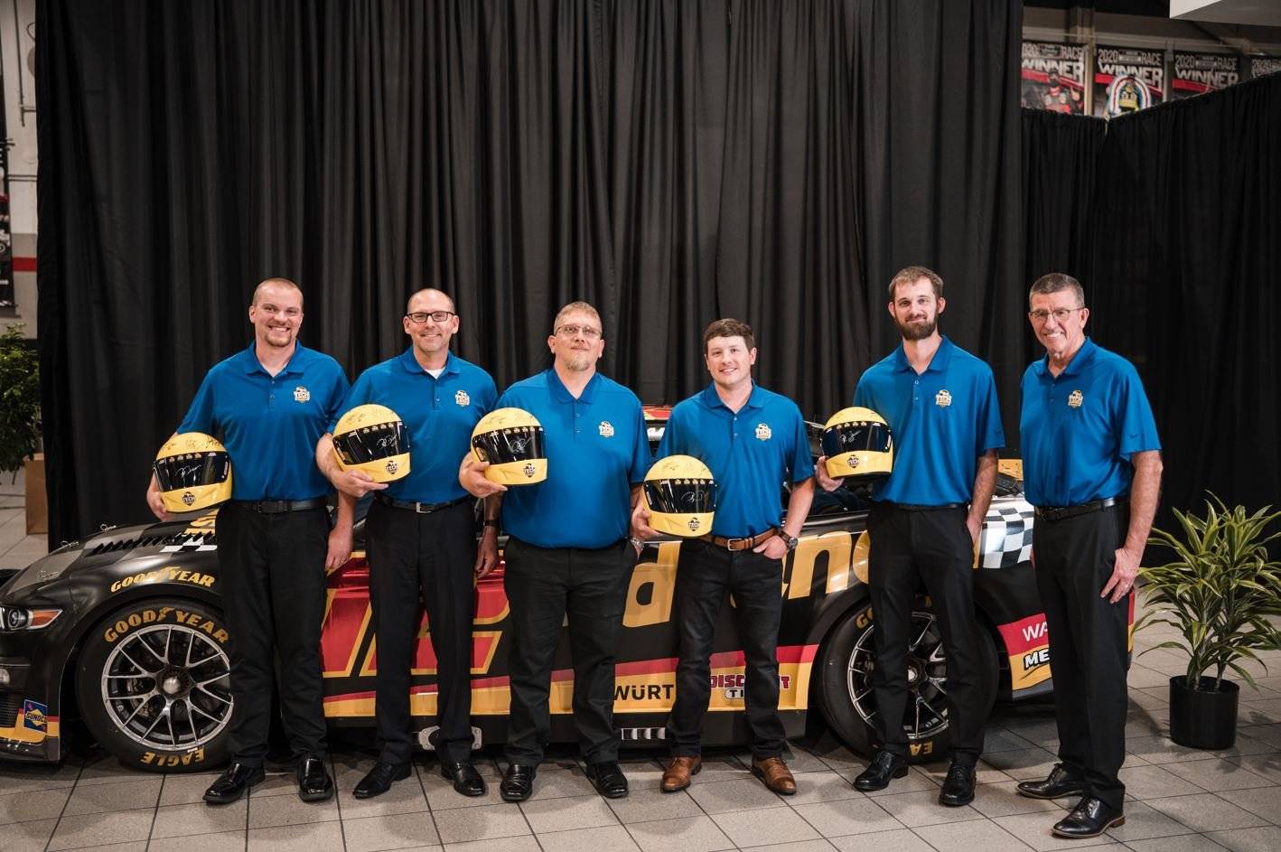 Six men posing in front of a race car holding helmets