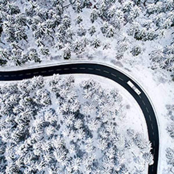 Snow-covered forest with a winding road and a white vehicle, aerial view.