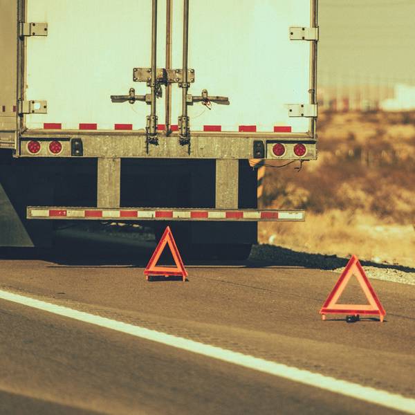 The back of a white semi-truck parked on the side of a road with two caution triangles set up behind it.