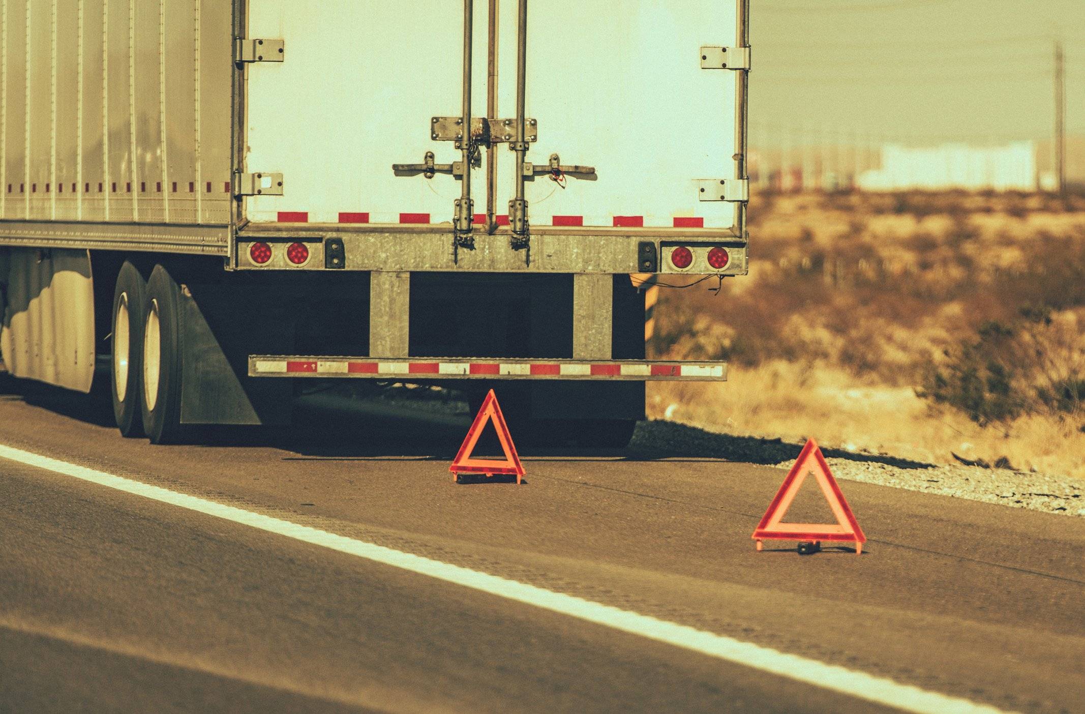 The back of a white semi-truck parked on the side of a road with two caution triangles set up behind it.