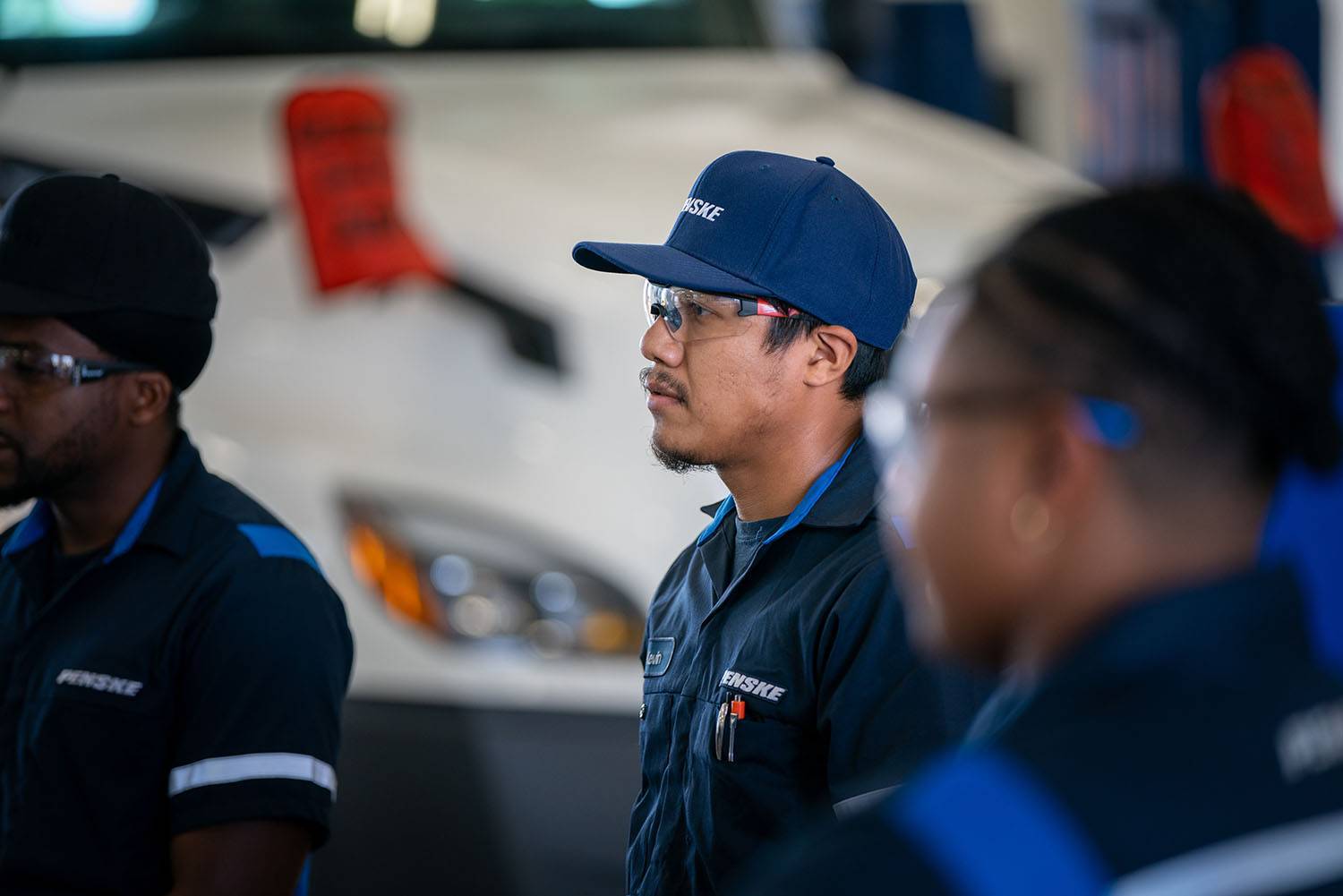 The profile of a diesel technician dressed in a blue Penske uniform and hat standing in front of a white semi-truck.