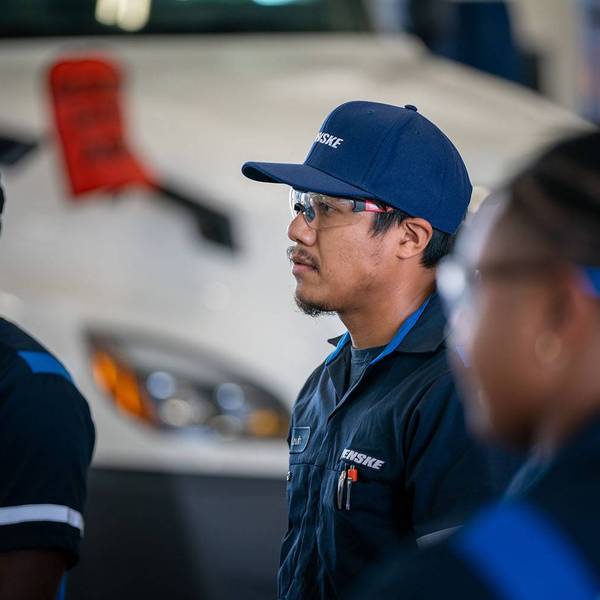 The profile of a diesel technician dressed in a blue Penske uniform and hat standing in front of a white semi-truck.