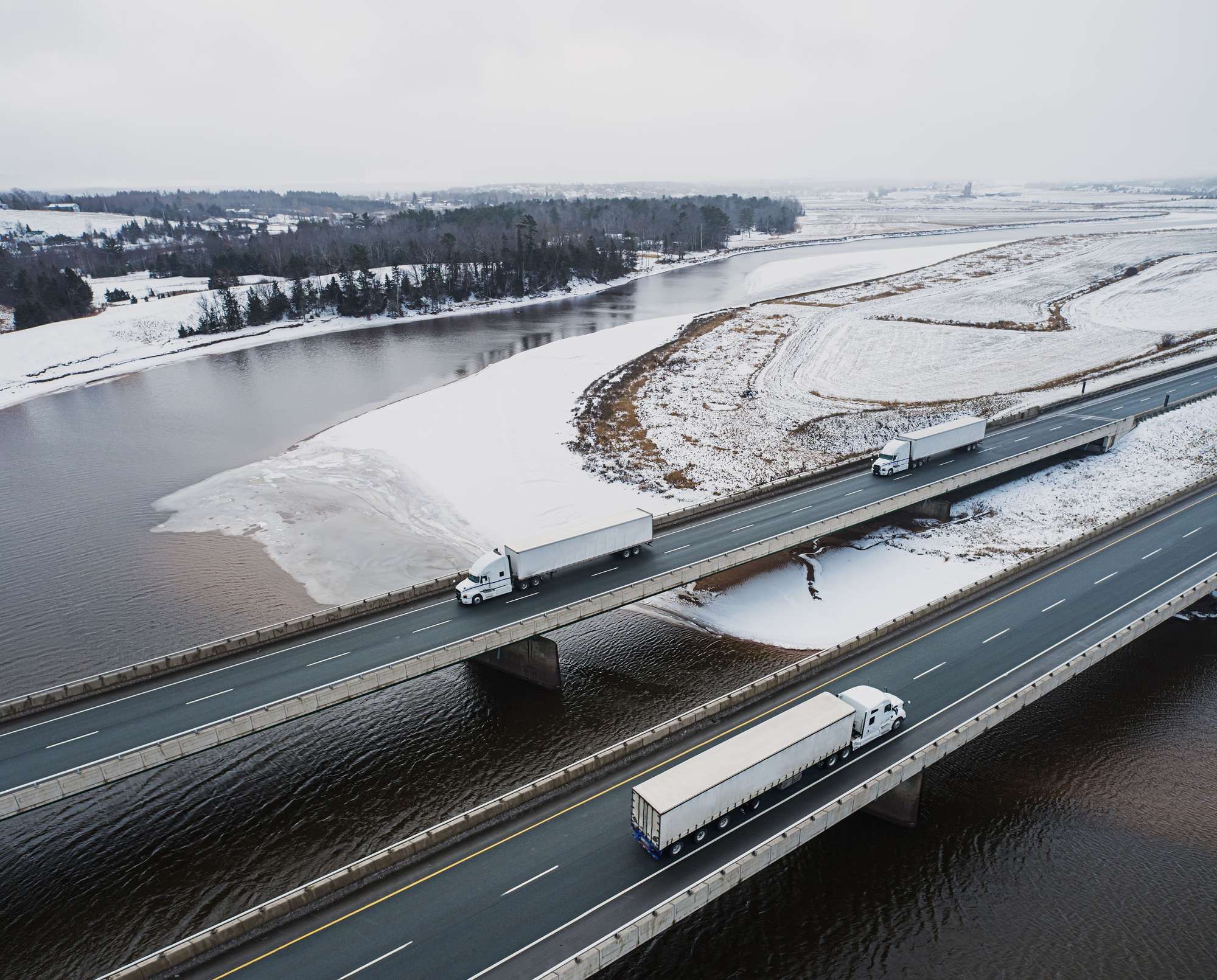 Three semi-trucks drive on a bridge across snowy land and water.