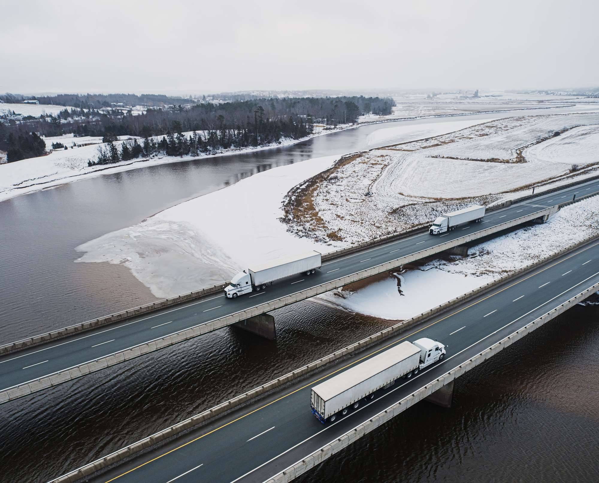 Three semi-trucks drive on a bridge across snowy land and water.