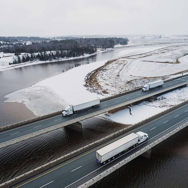 Three semi-trucks drive on a bridge across snowy land and water.