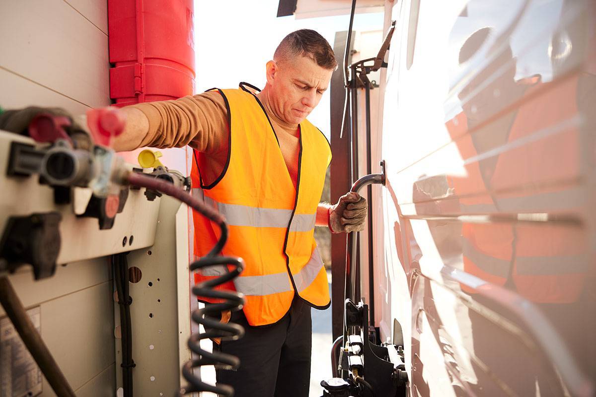 Truck driver in an orange vest and gloves examining semi-truck trailer brake coil for safety
