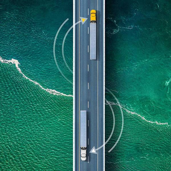 Two semi-trucks drive in opposite directions on a bridge over blue-green water.