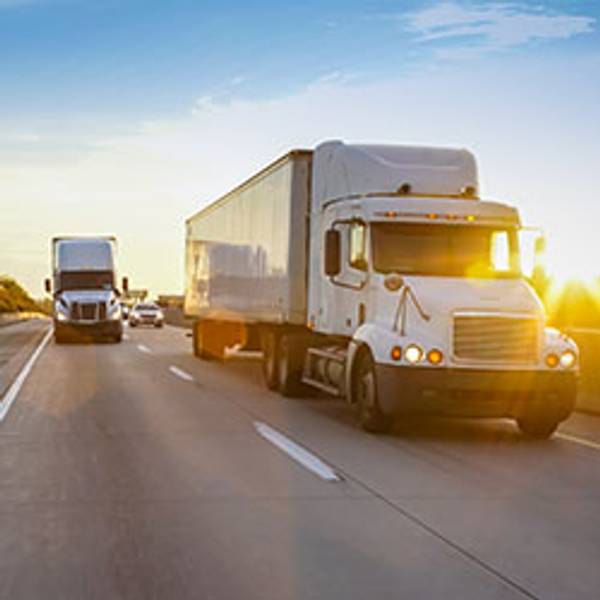 Two semi-trucks drive on a sunny highway at sunset.