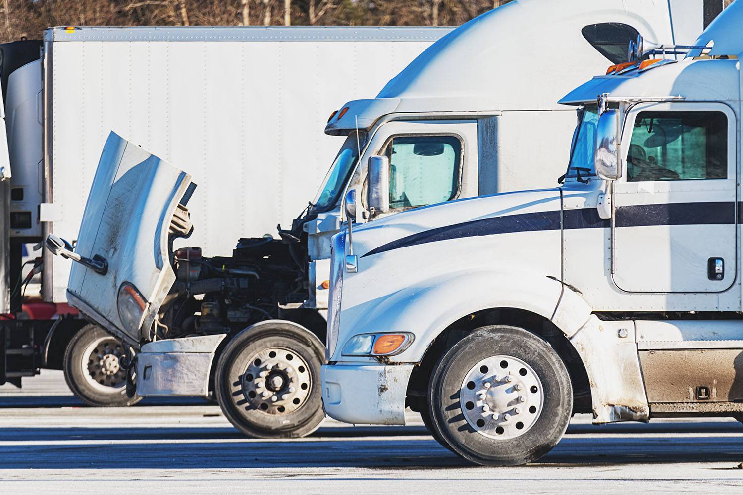 Two white semi-truck cabs sit parked with the front end of one open for maintenance.