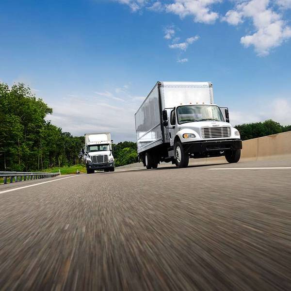 Two white trucks travel on a highway under a blue, cloudy sky.