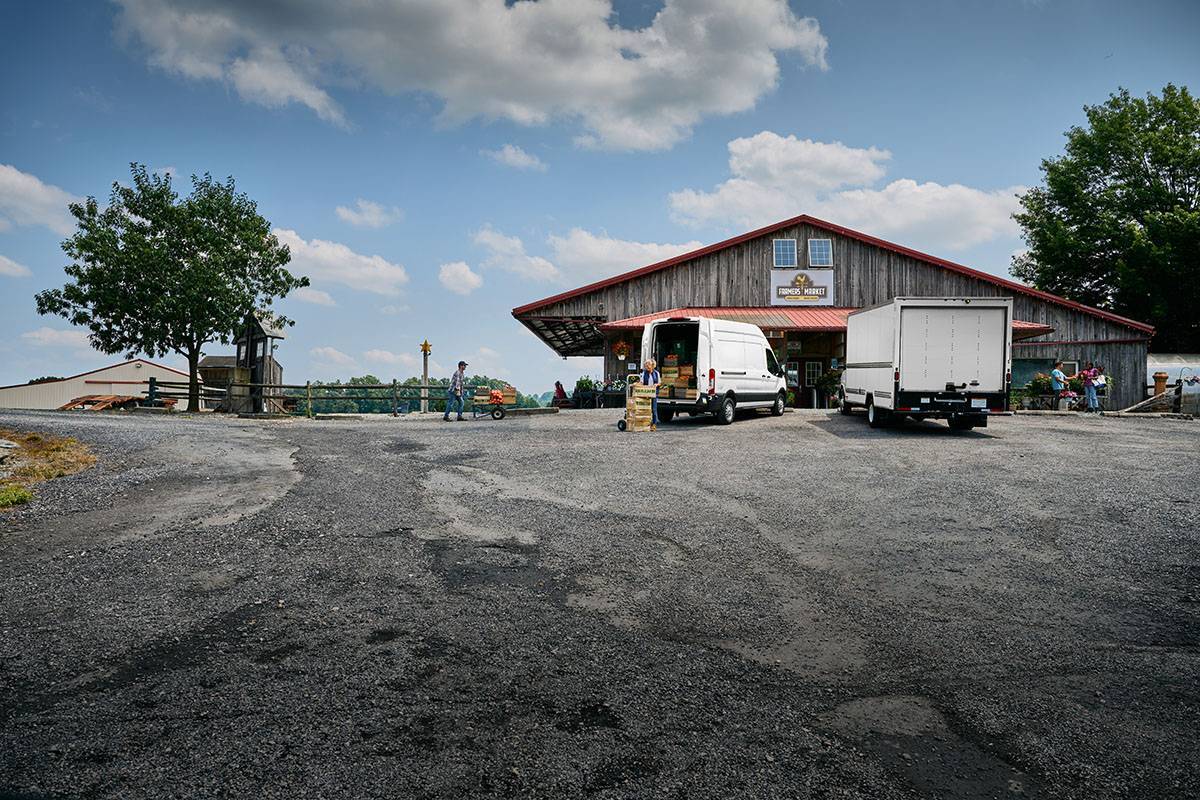 Two white trucks unload cargo outside of a farmer's market under a blue sky.