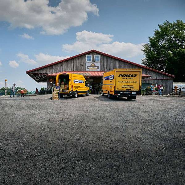Two yellow Penske trucks unload boxes outside a brown barn farmer's market