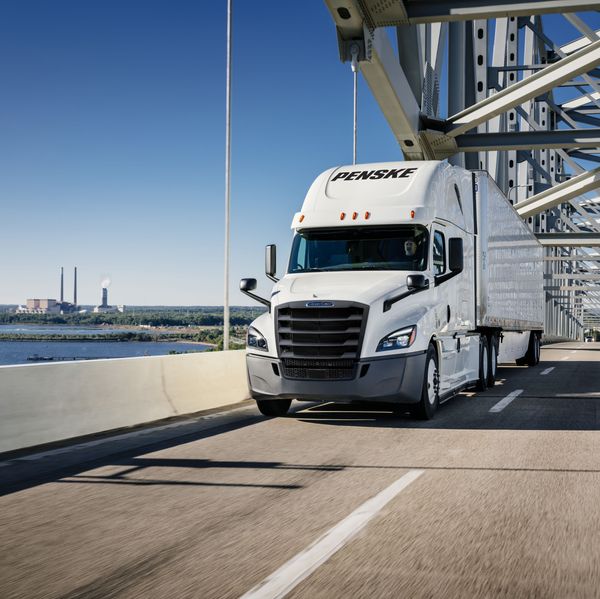 White Penske semi truck drives over a bridge next to water.