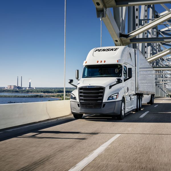 White Penske semi truck drives over a bridge next to water.