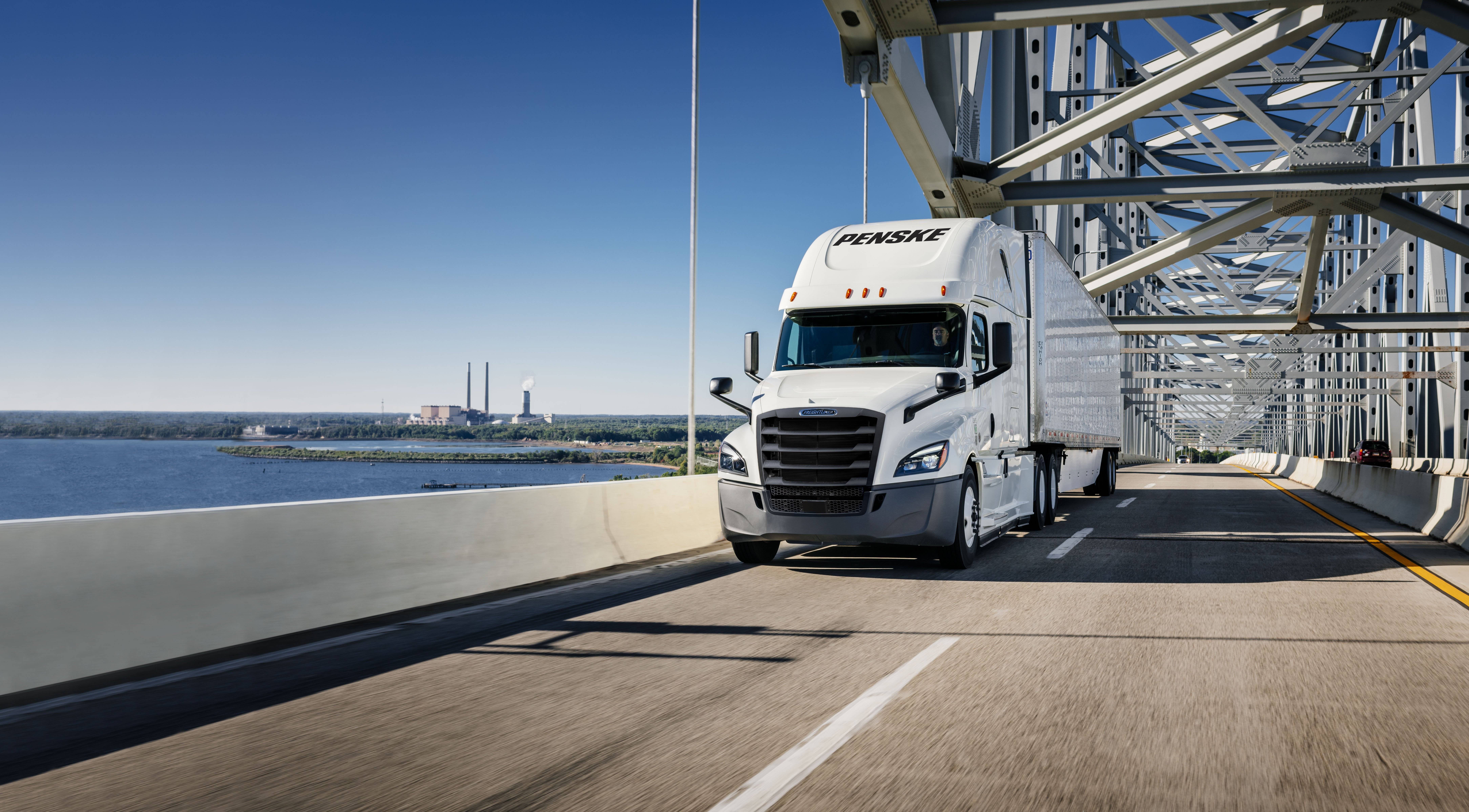 White Penske semi truck drives over a bridge next to water.
