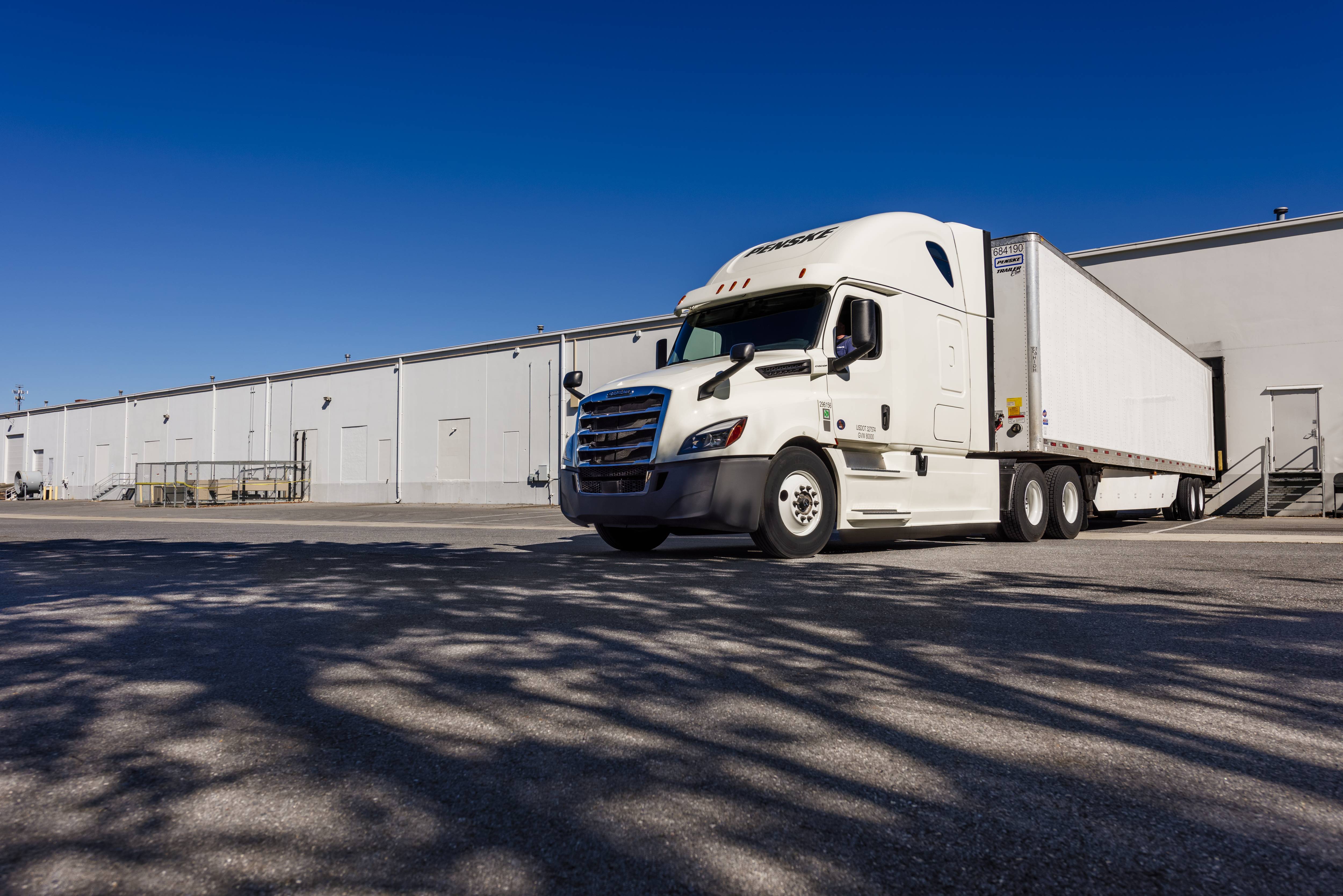 White Penske semi-truck is backed in to a loading dock in a warehouse parking lot.