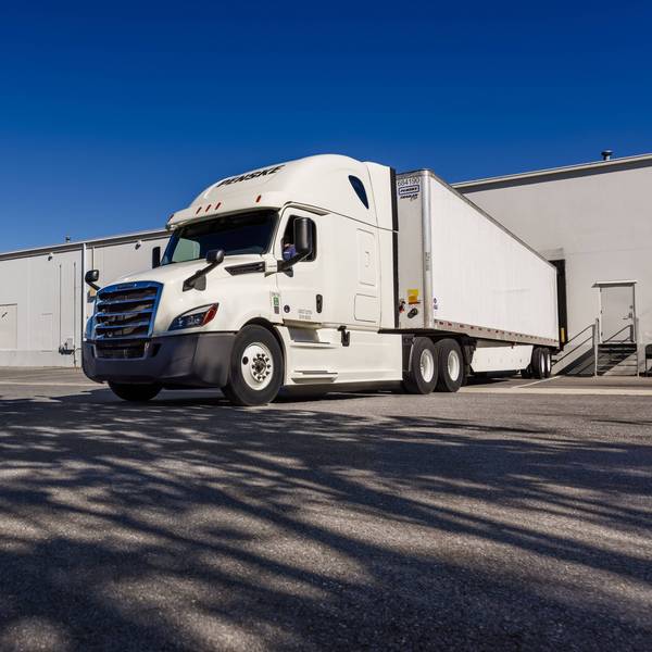 White Penske semi-truck is backed in to a loading dock in a warehouse parking lot.