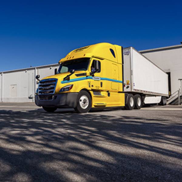 Yellow semi-truck with trailer parked by a warehouse under a clear blue sky.